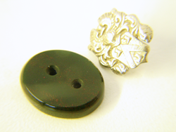 A finished oval flat topped Bloodstone cabochon with 2 holes in it sitting next to the white gold emblem.