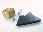 Photo of a square ring, a Diamond in a gold tube, a masonic emblem, and a piece of rough black Jade.